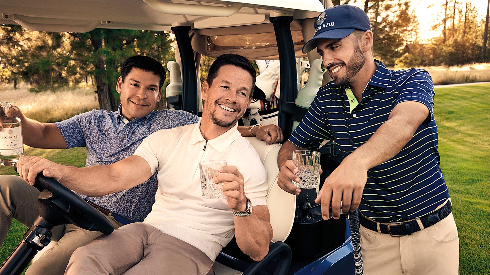 Mark Wahlberg, Aron Marquez and Abraham Ancer sit and stand in and around a golf cart with a bottle and glasses of their tequila, Flecha Azul.